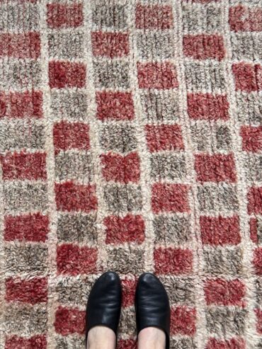 Alfombra vintage marroquí con diseño de cuadros en rojo, beige y marrón, tejida a mano en lana gruesa, colocada sobre suelo claro.