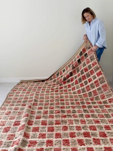 Alfombra vintage marroquí con diseño de cuadros en rojo, beige y marrón, tejida a mano en lana gruesa, colocada sobre suelo claro.
