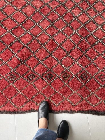 Alfombra bereber vintage en rojo intenso con patrón de rombos en crema y negro, tejida a mano en lana gruesa, sobre suelo claro