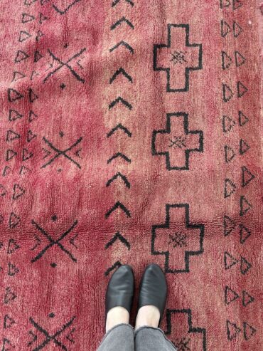 Alfombra vintage marroquí roja tejida a mano en lana gruesa, con motivos geométricos en un extremo, colocada sobre suelo claro.