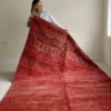 Alfombra vintage marroquí roja tejida a mano en lana gruesa, con motivos geométricos en un extremo, colocada sobre suelo claro.