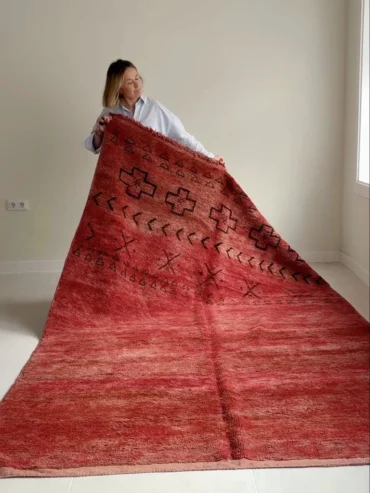 Alfombra vintage marroquí roja tejida a mano en lana gruesa, con motivos geométricos en un extremo, colocada sobre suelo claro.