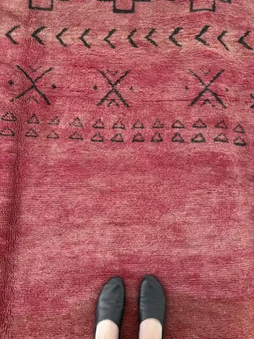 Alfombra vintage marroquí roja tejida a mano en lana gruesa, con motivos geométricos en un extremo, colocada sobre suelo claro.